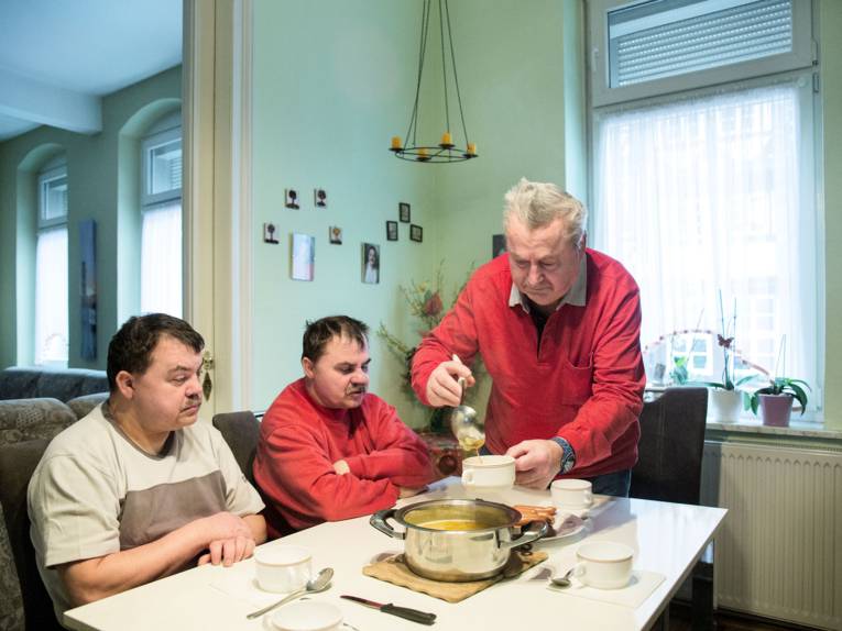 Zwei Männer sitzen an einem Tisch, ein dritter Mann schenkt ihnen Suppe ein.